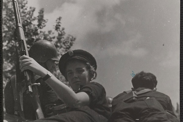 Portrait d'une jeune femme en uniforme, allongée sur un matelas, entre deux soldats. Armée, elle tourne le dos à ses compagnons et regarde le photographe. [1936-1939]. (Mémoires d’Humanité / Archives départementales de la Seine-Saint-Denis)